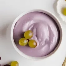 Fotografia em tons de branco e roxo com um pote branco ao centro. Dentro do pote existe uma receita de mousse de uva de cor roxa com algumas uvas verdes de enfeite.