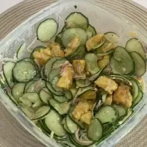 Fotografia em tons de bege com uma travessa de vidro ao centro. Dentro da travessa existe uma receita de salada de pepino com frango, cebola roxa e molho