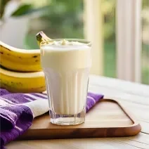 Fotografia em tons de roxo e amarelo com um copo de vidro ao centro. Dentro do copo existe um shake de coco, batata doce e banana de cor branca. Ao fundo existe duas bananas