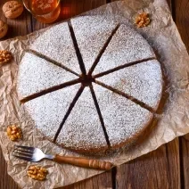 Foto em tons de marrom da receita de bolo alemão de mel servida cortada em 8 pedaços polvilhada com açúcar de confeiteiro, bolo em cima de uma mesa de madeira com nozes ao lado