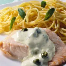 Uma mesa azul com bolinhas brancas vista de cima, um prato quadrado branco com macarrão espaguete, um pedaço de salmão com molho branco por cima.