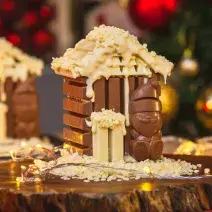 Imagem da receita de Casinha de KitKat Papai Noel, montada num formato miniatura de uma casinha de chocolate em tons marrom e branco, com um Papai Noel de chocolate à frente, sobre uma tábua de madeira e com decorações de natal ao redor