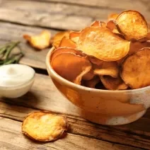 Fotografia em tons de marrom com um prato fundo de cerâmica ao centro. Em cima do prato existe uma porção de chips de batata doce feitos na Air Fryer