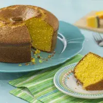 Fotografia em tons de azul de uma bancada azul com um prato azul, sobre ele um bolo de abóbora. Ao lado um paninho verde listrado com um prato redondo pequeno com uma fatia do bolo. Ao fundo uma tábua com uma fatia de abóbora.