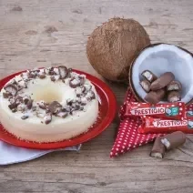 Foto em tons de vermelho e marrom da receita de pudim de prestígio servida em um prato de porcelana vermelho com panos brancos embaixo. Ao lado, um coco cortado ao meio com prestígios dentro e mais embalagens a frente, além de um pano vermelho com bolas