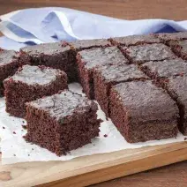 16 pedaços de bolo de chocolate com calda em cima de uma toalha branca que está em cima de uma tábua de madeira. Todos do mesmo tamanho