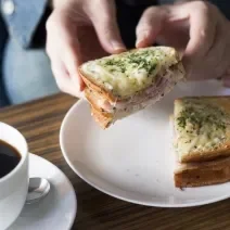 Fotografia de um sanduíche aberto com uma moça segurando a metade do lanche, que tem presunto, lombinho defumado, molho e queijo gruyère gratinado por cima. O sanduíche está em um prato raso branco de vidro ao lado de uma xícara branca de café cheia.
