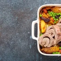 Fotografia vsita de cima de um refratário quadrado branco com vegetais e um lombo assado em fatias, recheado com farofa. o refratário está sobre uma bancada escura com detalhes azuis.