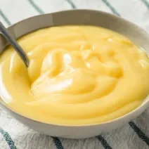 Foto aproximada de um pote redondo com um creme amarelo dentro e uma colher, tudo sobre um tecido de cor bege com listras verdes