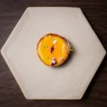 Fotografia em tons de cinza com uma cheesecake amarela ao centro. Por cima da cheesecake existe uma calda de bergamota, caramelo e flores comestíveis.