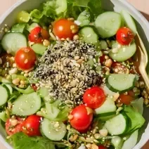 Fotografia mostra uma salada verde com tomate, pepino, grãos, sementes e um pouco de gergelim preto espalhado pela salada