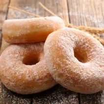 Fotografia de três rosquinhas, uma apoiada na outra, passadas no açúcar de confeiteiro, sobre uma mesa de madeira.