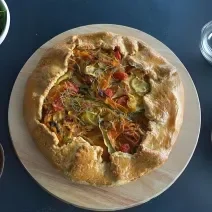 Fotografia em tons de marrom com uma torta rústica de legumes ao centro. Dentro da torta existe fatias de abobrinha, cenoura, alho poró e tomates.