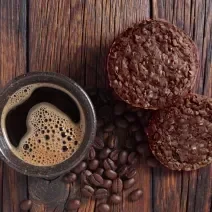 Foto vista de cima da receita de cookies de aveia e café, servidos em uma bancada de madeira, ao lado de uma xícara de café preparada com alguns grãos de café espalhados