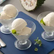 Fotografia em tons de branco e azul de uma bancada, ao centro uma tábua azul, com três taças com duas bolas de sorvete de graviola cada. Ao fundo, uma graviola cortada ao meio.