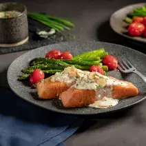 Foto de um prato escuro com a receita de salmão grelhado, acompanhado de aspargos e tomates cereja. Ao fundo há outro prato aparecendo, alguns aspargos um copo preto pequeno com molho dentro