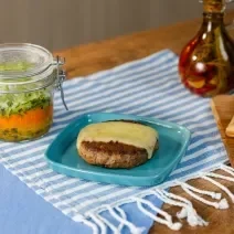 Fotografia em tons de azul com um prato azul ao centro. Em cima do prato existe uma receita de hambúrguer no prato acompanhada por uma fatia de queijo muçarela e uma salada no pote feita com alface e cenoura