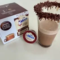 Fotografia de uma xícara de vidro com uma bebida de dolce gusto brigadeiro, com brigadeiro e granulado na borda. Ao lado tem uma cápsula de dolce gusto brigadeiro e a caixa com as cápsulas.