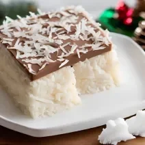 Foto da receita de prestígio na travessa, servido em fatia com uma camada de creme de coco coberta com ganache de chocolate e decorado com coco ralado, sobre uma mesa decorada para o natal