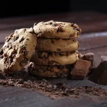 Fotografia de quatro cookies com gotas de chocolate, um sobre o outro. Encostado neles tem mais um cookie, ao lado de pedaços de chocolate, sobre uma mesa de madeira escura.