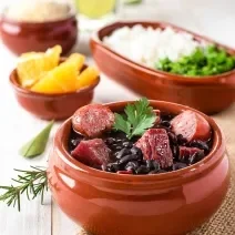 Fotografia de um recipiente médio marrom clássico com uma feijoada com caldo de bacon, linguiça e bacon com uma folha de salsinha. Ao fundo tem outros recipientes iguais, porém menores, com laranja, arroz, couve e farofa.