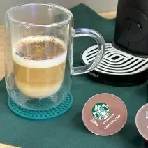 Foto da receita de Cappuccino Canela. Observa-se um copo transparente com o cappucino dentro, polvilhado de canela e chocolate em pó. Ao lado direito, cápsulas de Starbucks.
