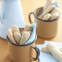 Fotografia em tons de azul em uma mesa de madeira com um paninho azul claro com bolinhas brancas, ao centro uma xícara bege com os biscoitinhos nutritivos. Ao fundo, uma jarra azul claro.