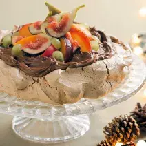 fotografia tirada de frente, contém um suporte para servir sobremesas transparente por cima um merengue coberto de chocolate com frutas para decorar.