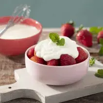 Fotografia em tons de vermelho em uma bancada de madeira clara, uma tábua de madeira branca, um potinho rosa claro com morangos dentro cobertos com creme de leite fresco. Ao fundo, um potinho vermelho com o creme de leite fresco dentro dele.