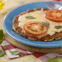 fotografia em tons de azul, vermelho e marrom de uma bancada marrom vista de cima, contém uma tábua redonda, um prato redondo azul com uma mini pizza coberta com queijo, duas fatias de tomate e manjericão e ao lado um garfo para servir.