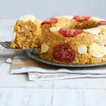 Fotografia em tons de branco e amarelo de uma bancada vista de frente, um pano branco e acima uma travessa com o cuscuz e rodelas de tomates. Ao lado uma espátula com uma fatia.