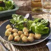 Fotografia em tons de azul e verde em uma bancada de madeira escura, um pano azul, um prato redondo raso branco com as vieiras acompanhadas de salada verde.