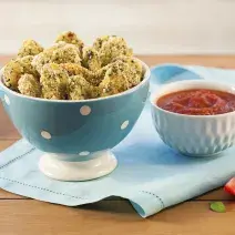 Fotografia em tons de marrom e azul claro de uma mesa de madeira vista de frente, contém um pano cor azul claro e por cima um recipiente redondo cor azul com bolinhas brancas com pedaços de nuggets, ao lado um pote branco com molho de tomate.