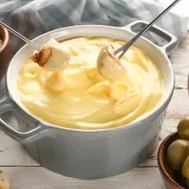 Fotografia em tons de branco com uma panela de fondue ao centro. Dentro da panela existe um fondue de queijo. Dois pedaços de pão estão sendo mergulhados no fondue de queijo. Ao fundo existe recipientes com azeitona e pedaços de pão.
