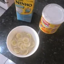 Fotografia de um recipiente branco fundo com um creme de farinha láctea com banana fatiada sobre uma bancada preta.