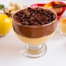 Fotografia de uma sobremesa de maracujá, chocolate e biscoito em uma taça, que está sobre uma mesa branca. Ao fundo, diversas opções de mousse de maracujá, a fruta inteira e uma tábua de madeira clara.