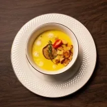 Fotografia em tons de marrom com um prato de sopa branco ao centro. Dentro do prato existe um creme de cor amarela feito com mandioquinhas acompanhado com croutons, cogumelos e tomate cereja.