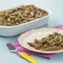 Fotografia em tons de azul e rosa em uma bancada de madeira azul, um guardanapo de pano rosa com bolinhas brancas, um refratário branco retangular com o picadinho de carne com quiabo. Ao lado, um prato rosa redondo com uma porção do picadinho e arroz.