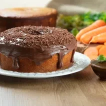 Fotografia de dois bolos de cenoura, um sem e o outro com cobertura de chocolate cremoso e granulado, apoiado em um prato branco. Ao lado tem mais cobertura em um recipiente pequeno de vidro, e ao fundo tem cenouras inteiras e alguns pedaços.