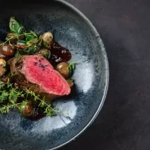 Fotografia vista de cima de um de assado ao molho ferrugem delicioso feito com Filé mignon temperado, com molho de vinho tinto e champignons. A receita está em um prato fundo de tom azul escuro, sobre uma mesa de cimento queimado escuro.