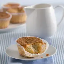 Fotografia em tons de azul em uma mesa de madeira com uma toalha listrada azul, um paninho azul, um pratinho de sobremesa branco com o muffin de banana com iogurte. Ao fundo, um bule branco com chá dentro.