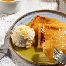 Foto de um prato em tom claro com a receita de Crepe Suzette, servido com calda amarela e uma bola de sorvete. O prato está em uma bancada clara, com. um pote de calda amarela e um guardanapo ao lado.