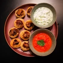 Fotografia vista de cima de oito pani puris de berinjela, e ao lado tem molhos de iogurte e pimentão em recipientes separados. Os pani puris e os recipientes estão apoiados em um prato de tom vermelho, o qual está apoiado sobre uma bancada preta.