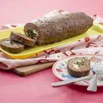 Fotografia em tons de rosa em uma mesa rosa com um pano rosa desenhado e um recipiente retangular amarelo com o bolo de carne com espinafre em cima dele. Ao lado, um prato colorido com uma fatia do bolo e um garfo e uma faca apoiados sobre ele.