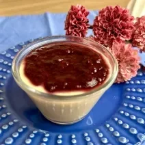 Fotografia em tons de azul e rosa com um pote transparente ao centro. Dentro do pote existe uma receita de flan de mamão coberto com geleia de frutas.