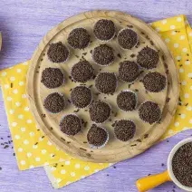 Fotografia em tons de marrom, amarelo e lilás, com bancada lilás vista de cima, ao centro peça de madeira com brigadeiros sobre guardanapo amarelo com bolinhas brancas, ao lado potinho com granulado, um brigadeiro e e duas bananas.