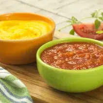 Fotografia em tons de verde, amarelo e vermelho com dois potes ao centro, sendo um amarelo com creme amarelo e um verde com um creme de tomate, ambos sobre peça de madeira, ao lado guardanapo e fatias de tomate, tudo sobre bancada de madeira.