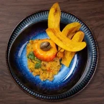 Fotografia em tons de marrom com um prato preto ao centro. Em cima do prato existe uma receita de ceviche de caju com castanhas e chips de banana da terra