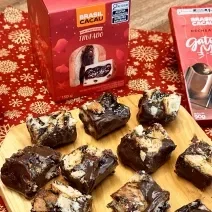 Foto da receita de palha italiana de mini panettone brasil cacau, servida em pedaços pequenos sobre uma tábua de madeira, em uma bancada decorada com itens natalinos e produtos da marca brasil cacau ao redor