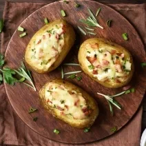 Fotografia vista de cima de três batatas assadas e recheadas com requeijão, salsinha e bacon por cima. As batatas estão sobre uma tábua de madeira redonda, e a tábua está sobre uma mesa de madeira escura. Ao redor tem mais batatas, queijo a salsinha.